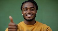 Young African American man with braided hair wearing yellow sweater showing thumbs up gesture with bright genuine smile against green background.