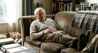 Senior Caucasian man relaxing on comfortable sofa in cozy living room with bookshelves, enjoying peaceful retirement lifestyle at home.