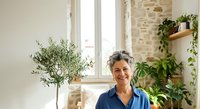 Mature woman with gray hair and Mediterranean ethnicity smiling at camera in bright, plant-filled home interior with natural light from windows and potted greenery surrounding her.