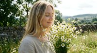 Young blonde woman smelling white wildflowers in countryside meadow with peaceful expression enjoying nature outdoors during golden hour summer day.