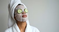 Young woman enjoying spa treatment with cucumber slices on eyes and facial mask wearing white bathrobe and towel for relaxation and skincare routine.