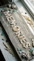 Rustic driftwood plank with seashells, sea glass, and sand arranged into letters, decorated with dried grass and pebbles on a windowsill.