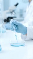 Scientist in white lab coat and blue latex gloves pipetting blue liquid sample into petri dish in sterile laboratory setting with microscope in background.