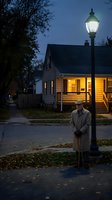 Elderly Caucasian man in beige coat standing under vintage street lamp at dusk in suburban neighborhood with warm house lights and autumn leaves on sidewalk.