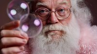 Senior Caucasian man with white beard and round glasses blowing soap bubbles against dark background, creating magical festive atmosphere.