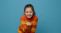 Happy young girl in colorful striped sweater smiling warmly while hugging herself on blue background. Cheerful child expressing joy and self-love concept.