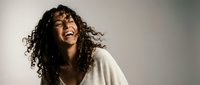 Young African American woman with curly hair laughing joyfully in white shirt against neutral background for lifestyle and wellness content.