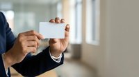 Businessman in dark suit holding blank white business card in modern office environment with natural light from windows.