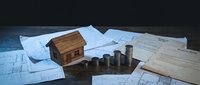 Wooden house model with stacked coins on architectural blueprints and documents for real estate investment planning and property development concepts.