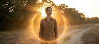 Young African American man standing on rural dirt road during golden hour sunset with warm sunlight creating dramatic backlighting through trees in peaceful countryside setting.