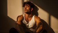 Young athletic man in white tank top sitting against wall with dramatic shadow play and warm golden light creating moody atmosphere.