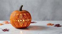 Carved pumpkin with intricate geometric zigzag pattern glowing from within, surrounded by autumn leaves on white surface for Halloween decoration.
