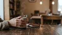 Elderly hands holding steaming coffee cup on wooden table in cozy cafe interior with warm lighting and blurred background for relaxation concepts.