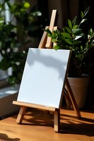 Wooden easel with blank white canvas in natural sunlight, green plant leaves creating soft shadows, artistic workspace setup on wooden table surface.