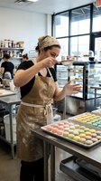 Female pastry chef in apron dusting powder over colorful macarons on trays in a professional bakery kitchen.
