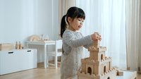 Young Asian girl in striped shirt and apron building wooden block castle in bright playroom, focused on creative construction play and learning.