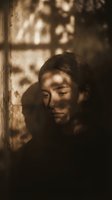 Young Asian man in contemplative mood with warm sunlight and shadow patterns creating atmospheric portrait for lifestyle and wellness content.
