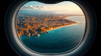 Aerial coastal landscape view through airplane window, dramatic cliffs meeting turquoise ocean waters at sunset, mountainous shoreline with golden light illuminating rugged terrain.