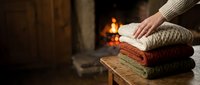 Cozy stack of folded knit sweaters on wooden table with fireplace glow in background, perfect for autumn and winter lifestyle, home decor, and seasonal marketing campaigns.