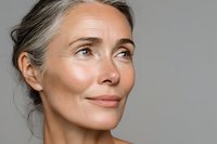 Mature woman with natural gray hair and glowing skin looking upward with serene expression against neutral gray background.