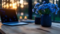 Cozy workspace with laptop and blue flowers in vase on wooden table at sunset, warm bokeh lights through window create peaceful home office atmosphere.