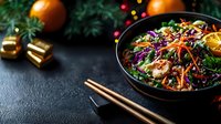 Asian noodle salad with vegetables, sesame seeds and citrus in black bowl on dark background with Christmas decorations and chopsticks.