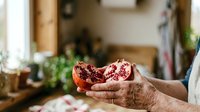 Senior woman with wrinkled hands holding open pomegranate with red seeds in bright kitchen, showing fresh fruit preparation and healthy eating lifestyle.