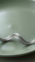 Strand of silver hair resting on pale green ceramic plate, creating elegant wave pattern against soft neutral background for beauty and wellness design projects.