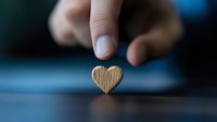 Small wooden heart shape balanced on dark surface with finger reaching from above, creating minimalist composition with shallow depth of field and soft focus.
