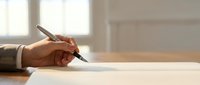 Person signing document with fountain pen at wooden desk in bright office setting with window light.