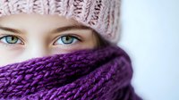 Close up portrait showing bright blue eyes beneath pink knitted winter beanie hat and wrapped in purple chunky knit scarf, expressing warmth and winter fashion style.
