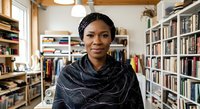 Young African woman with dark hair wearing a dark patterned scarf in a home library surrounded by bookshelves and natural light from windows, suitable for education and lifestyle content.