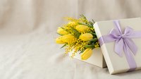 Yellow tulips and mimosa flowers with elegant gift box and purple ribbon on soft beige background for spring celebrations and special occasions.