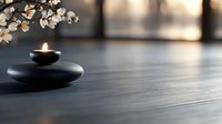 Zen spa composition with glowing candle on black stones and white cherry blossom branch against blurred background, creating peaceful atmosphere.