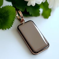 Rose gold rectangular pendant necklace charm with smooth polished surface against white background with green leaves and flowers, jewelry accessory.