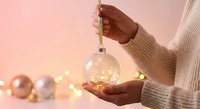 Female hands in cozy knit sweater holding glowing Christmas ornament with warm fairy lights and festive decorations on soft pink background for holiday design.