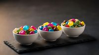 Three white ceramic bowls filled with colorful round candies arranged on dark slate board against moody background for confectionery and dessert concepts.