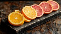 Fresh orange and pink grapefruit halves arranged in row on rustic wooden board, showing vibrant citrus flesh and segments in warm lighting.