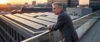 Mature businessman in dark suit standing on urban rooftop at sunset, leaning on glass railing overlooking city skyline with modern buildings and golden hour light.