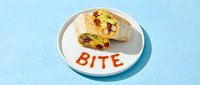 Breakfast burrito cut in half on a white plate with the word BITE written in red sauce, served on a light blue background.