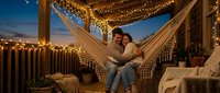 Couple relaxing in hammock on illuminated rooftop terrace at dusk, surrounded by string lights and potted plants for outdoor entertaining and romantic evening gatherings.