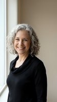 Confident mature woman with gray curly hair smiling in black sweater by window, professional portrait for business and lifestyle content.