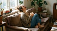 Young woman relaxing on cozy sofa with coffee cup in bright living room with plants. Comfortable home lifestyle moment for wellness and self-care content.