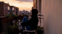 Young woman relaxing on urban apartment balcony during golden hour sunset with city skyline view and potted plants creating peaceful evening atmosphere.