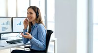 Happy young businesswoman wearing headset talking on phone at modern office desk with computer monitors showing charts and data analytics.