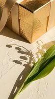 Elegant golden gift box with satin ribbon and lily of the valley flowers casting shadows on textured cream surface for luxury packaging and celebration.