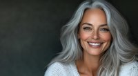Confident mature woman with silver gray hair and bright smile wearing white sweater against dark background, radiating natural beauty and positive aging concept.