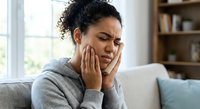 Young African American woman experiencing severe toothache pain holding jaw with both hands sitting on couch at home suffering from dental problems.