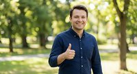 Happy Caucasian businessman in navy shirt giving thumbs up gesture outdoors in sunny park setting for positive business and success concepts.