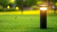Modern black garden bollard light illuminated at dusk on green lawn with soft bokeh background, outdoor landscape lighting design element.